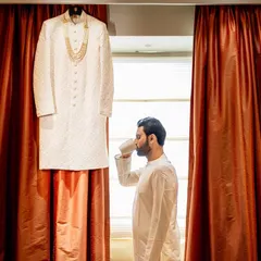 A creative shot of the groom enjoying a quiet cup of coffee before getting into his sherwani, which hangs in the background. A moment of calm before the celebration begins.