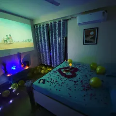 A full view of the bedroom decorated for a special occasion, with a heart of rose petals on the bed, balloons, and a movie playing on the projector screen.