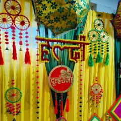 A close-up of a Haldi backdrop, showing the detailed floral work on the 'Haldi' sign and the 'Dulheraja' plaque, along with colourful tassels and hangings.