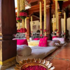 Details that honor tradition. A brass lotus bowl filled with fresh rose petals sits at the base of a wooden pillar, with jasmine and rose garlands hanging from the ceiling.