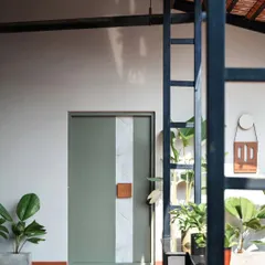 This perspective highlights the interplay of materials at the office entrance. The dark metal frame of the structure contrasts with the light walls and green door, while the natural light casts soft shadows, adding depth and character to the space.