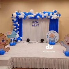 A stage setup for a Baptism or naming ceremony. This blue and white theme includes a balloon arch, a personalized sign, and cute baby character cutouts.