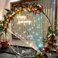 I design elegant backdrops like this anniversary ring for special celebrations. This setup features a large metal ring decorated with fresh roses and ferns, a custom 'Happy Anniversary' neon sign, and delicate fairy lights, creating a perfect photo opportunity.