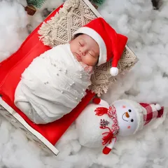 A simple and sweet Christmas portrait. A newborn in a Santa hat sleeps on a red sleigh, next to a friendly snowman prop on a bed of fluffy snow.