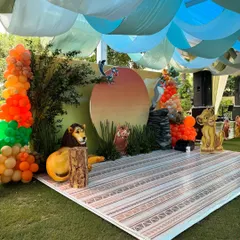 This outdoor Lion King party setup shows the complete stage with custom flooring that mimics an African textile pattern. The scene includes character cutouts of Mufasa, Simba, and Pumbaa, along with vibrant orange and green balloon columns.