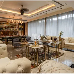 This elegant living area is designed for sophisticated entertaining, with a fully stocked bar and dining setup in the background. The tufted sofas, nested coffee tables, and sheer curtains create a layered look of luxury, with a view of the poolside just outside.