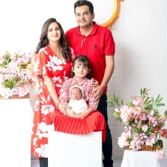 The complete family of four, beaming with joy. The older sister sits proudly with her new baby sibling, surrounded by her loving parents in a beautifully coordinated red-themed portrait.