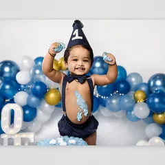 A triumphant birthday boy raises his frosting-covered hands in the air, celebrating his first birthday with a cake smash. The blue and gold balloon arch adds a festive touch to the studio session.