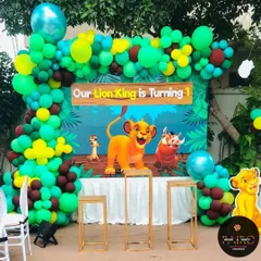 A vibrant Lion King theme for a first birthday party. This outdoor setup features a large backdrop with Simba and friends, framed by a jungle-inspired balloon arch in shades of green, yellow, and brown.