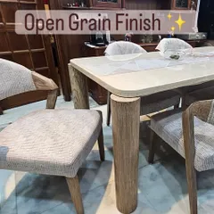 This dining set features a modern open-grain finish that highlights the natural beauty of the wood. The table has unique cylindrical legs, and the upholstered chairs have gracefully curved wooden armrests.