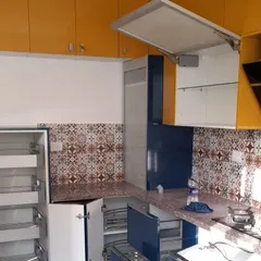This kitchen combines bold colors with functionality, featuring yellow upper cabinets and blue lower units. The design includes a lift-up cabinet door for easy access and patterned backsplash tiles that add a unique character.