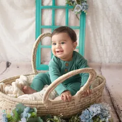 A happy six-month-old baby boy smiles from a wicker basket. The blue floral props and rustic window create a charming, cheerful scene.