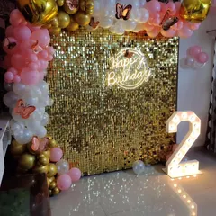 A beautiful second birthday decoration with a gold sequin wall. The backdrop is framed with a pink, white, and gold balloon arch with butterfly accents, a 'Happy Birthday' neon sign, and a large marquee number '2'.