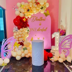 A "one-derful" butterfly-themed first birthday decoration. The setup uses a pink arch backdrop, a pastel yellow and red balloon garland, and large butterfly wing cutouts next to a light-up number '1'.