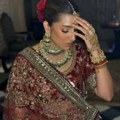 A candid moment of the bride, Ayushi, looking effortlessly stunning. Her makeup features warm tones and a subtle glow, complementing her deep red Sabyasachi lehenga and the intimate, regal setting of her wedding.