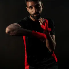 A close-up portrait of a fighter in a defensive stance. The red hand wraps indicate he is ready for striking drills, focusing on elbow blocks and head movement.