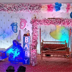 This baby shower decoration combines several popular elements. It has a moon prop with a Krishna theme, a floral swing, and 'It's a Boy/Girl' signs for a gender reveal.