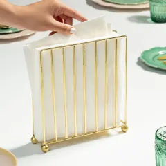 A styled shot of a gold napkin holder, shown in use on a set table. The human element of a hand reaching for a napkin makes the scene feel more dynamic and relatable.
