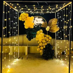 A stylish birthday party setup on a terrace. The main feature is a canopy frame decorated with curtains of fairy lights, leading to a black and gold balloon backdrop with a neon sign.