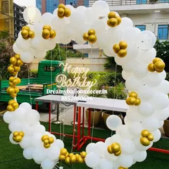 An elegant outdoor birthday setup on a lawn. The white and gold balloon ring with a 'Happy Birthday' neon sign looks beautiful in natural daylight.