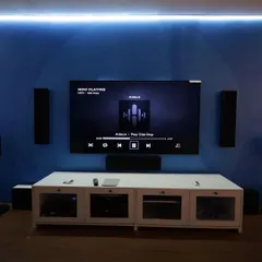A dedicated media room featuring a multi-speaker setup for surround sound, complemented by ambient blue backlighting to create an immersive viewing environment. The components are neatly organized in a white console below the screen.