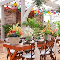 A dining table setup from the 'Bora Bora' event. The wooden table is adorned with tropical flower arrangements, jute-wrapped vases, and woven placemats, all under a canopy of colorful hanging lanterns.