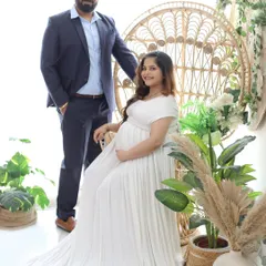 A classic boho couple portrait. The mom-to-be is seated on the peacock throne in a simple white gown, while her partner stands lovingly by her side.