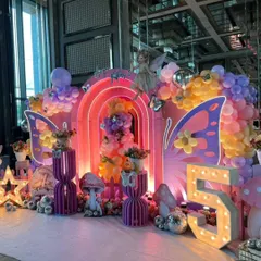 A vibrant and magical fairy-themed 5th birthday party. The decor is filled with color, featuring rainbow-arched backdrops, large butterfly wings, mushroom props, and a light-up number "5".