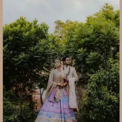 A regal portrait of a couple in exquisite designer outfits, standing amidst vibrant green trees. The bride's multi-colored lehenga by Tarun Tahiliani is a centerpiece of this elegant shot.