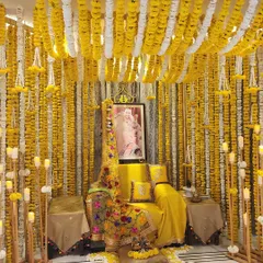 A stunning setup for a Satsang evening at home, bathed in auspicious yellow. The space is enveloped in curtains of yellow and white marigolds, with a comfortable seating area and the warm glow of many candles.