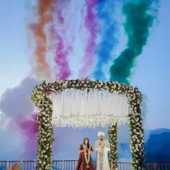 A moment of pure celebration as colorful smoke fills the sky behind the mountain mandap. This shot captures the festive energy of the Varmala ceremony, framed by the serene beauty of the hills.