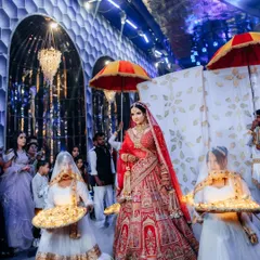 A royal bridal entry, with the bride flanked by attendants carrying decorative platters. The grandeur of the venue and her magnificent lehenga make for a majestic sight.