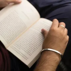 A man reading a book, wearing a silver cuff engraved with a name. This image captures the essence of the bracelet as a subtle, personal accessory for quiet moments.