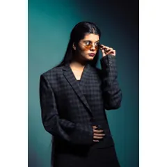 A studio shot of a model in a plaid blazer, adjusting her sunglasses. The teal background and professional lighting create a polished, high-fashion look suitable for a brand's lookbook or magazine feature.