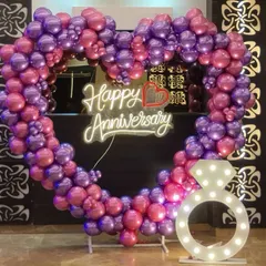 A romantic anniversary decoration featuring a heart-shaped balloon arch in purple and pink. A "Happy Anniversary" neon sign and a light-up ring prop complete this perfect photo spot.