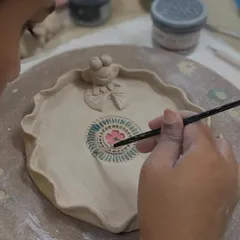 A student carefully paints a colorful mandala design onto a dish, complete with a little frog sculpture. This is part of our glazing masterclass.