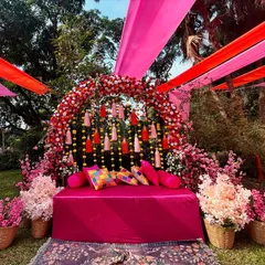 A lush, outdoor Mehendi setup with a floral arch in shades of pink and red, complemented by overhead drapes that add a canopy of color.