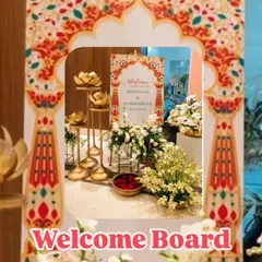 A close-up of a welcome area for a traditional wedding. The personalized welcome board is framed by an arch with classic Indian motifs, complemented by brass lotus candle stands and white floral arrangements.