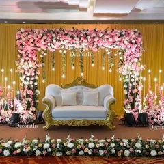 An elegant engagement stage setup. A classic white and gold sofa is framed by a beautiful arch of pink flowers, with hanging light bulbs and candle stands adding a warm, romantic glow.