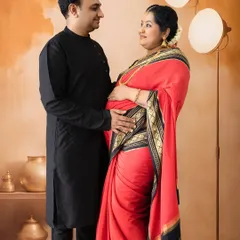 A classic and warm maternity portrait of a couple in traditional attire. The simple, earthy-toned backdrop and soft lighting create an intimate and loving atmosphere.