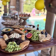 A sun-drenched poolside grazing setup perfect for a daytime celebration. The table is laden with multiple wooden boards featuring crackers, dips, fresh grapes, and other savory snacks, creating a relaxed yet luxurious atmosphere.