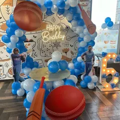 A cricket-themed 6th birthday party for a young sports fan. The setup includes giant cricket bat and ball props, player cutouts, and a blue and white balloon arch.