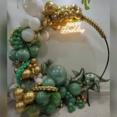 A roaring good time with this dinosaur-themed birthday setup. This impressive organic balloon ring in shades of green and gold is accented with tropical leaves and a large triceratops figure, with a "Happy Birthday" neon sign glowing in the center.