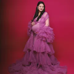 A smiling mother-to-be in a flowing, tiered purple gown. The studio lighting is set to beautifully illuminate her and the rich color of the dress against a solid magenta background.