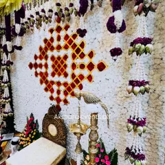 A detailed view of a Haldi or Sreemantham backdrop. The design features a wall of white flowers with a traditional pattern in red, and is adorned with hanging lotus bud garlands.