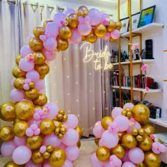 A chic and modern bridal shower decoration featuring a gold metal frame. The asymmetrical balloon arch in pink and gold, paired with a "Bride to be" neon sign and fairy lights, creates a perfect, picture-ready spot.