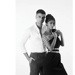 A full-length black and white portrait showing the couple's modern styling. The contrast between his simple shirt and her elegant dress with a coin belt creates a balanced and fashionable composition for this editorial.