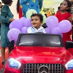 A happy little client enjoying his grand entrance. He is seated in a bright red toy Mercedes, decorated with a garland of purple balloons, ready to make his big debut at the party.