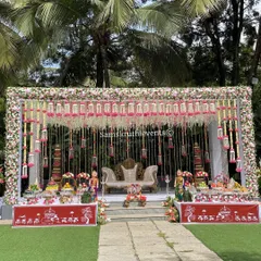 I love creating stunning outdoor wedding stages like this one. This setup for a traditional South Indian ceremony features hanging floral garlands, elegant seating, and a beautiful display of sweets and offerings, all set against a natural green backdrop.