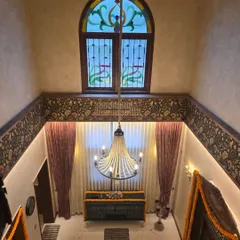 A view from the upper level of the Veerabhavana Villa, looking down into a double-height living space. A grand chandelier hangs below a stained-glass arched window, showcasing the home's blend of traditional details and dramatic scale.